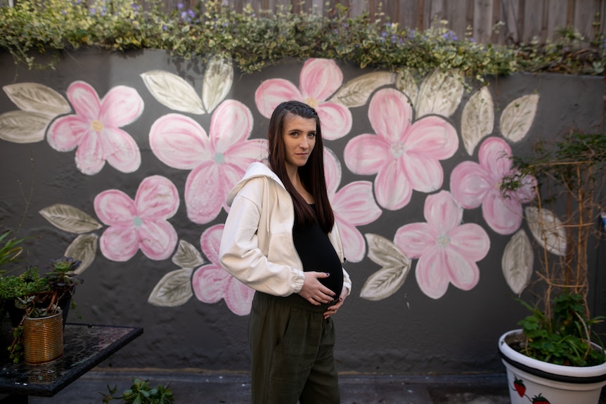 Alysha stands with her hands on her stomach looking at the camera with a neutral expression. Behind her is a large flower mural.
