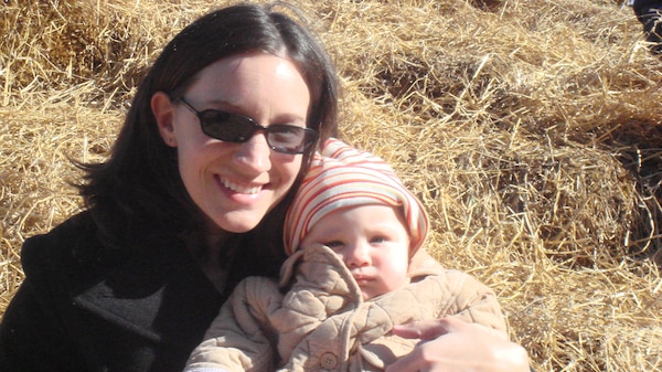 A dark-haired woman wearing sunglasses and holding a small baby wearing a jacket and beanie.