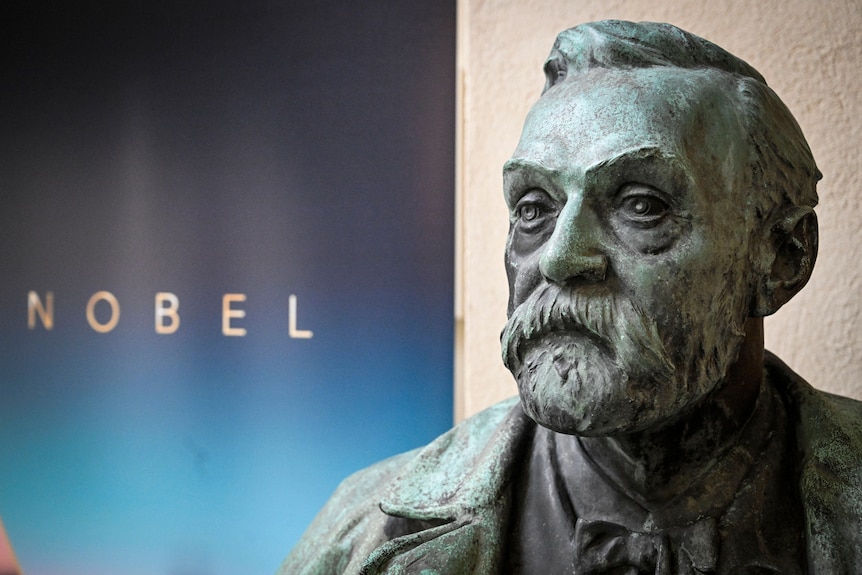 A bust of a bearded man sits in front of a wall-mounted sign saying NOBEL.