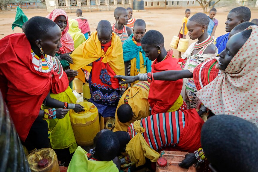 a dozen women in bright traditional african dress gather around plastic water vessels