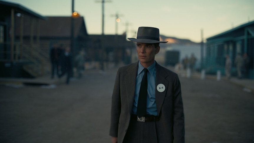 White man wearing a blue shirt, dark grey suit, hat and tie walks along an empty street at twilight.