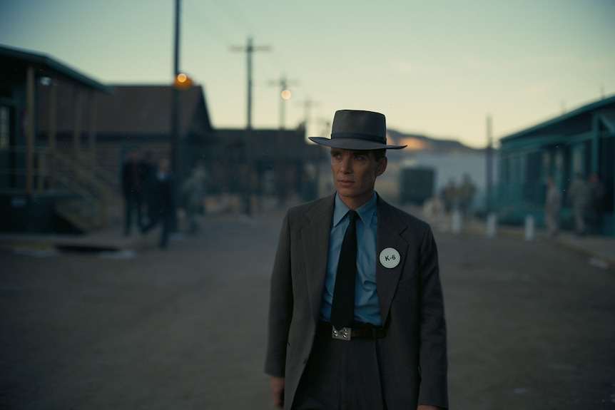 White man wearing a blue shirt, dark grey suit, hat and tie walks along an empty street at twilight.