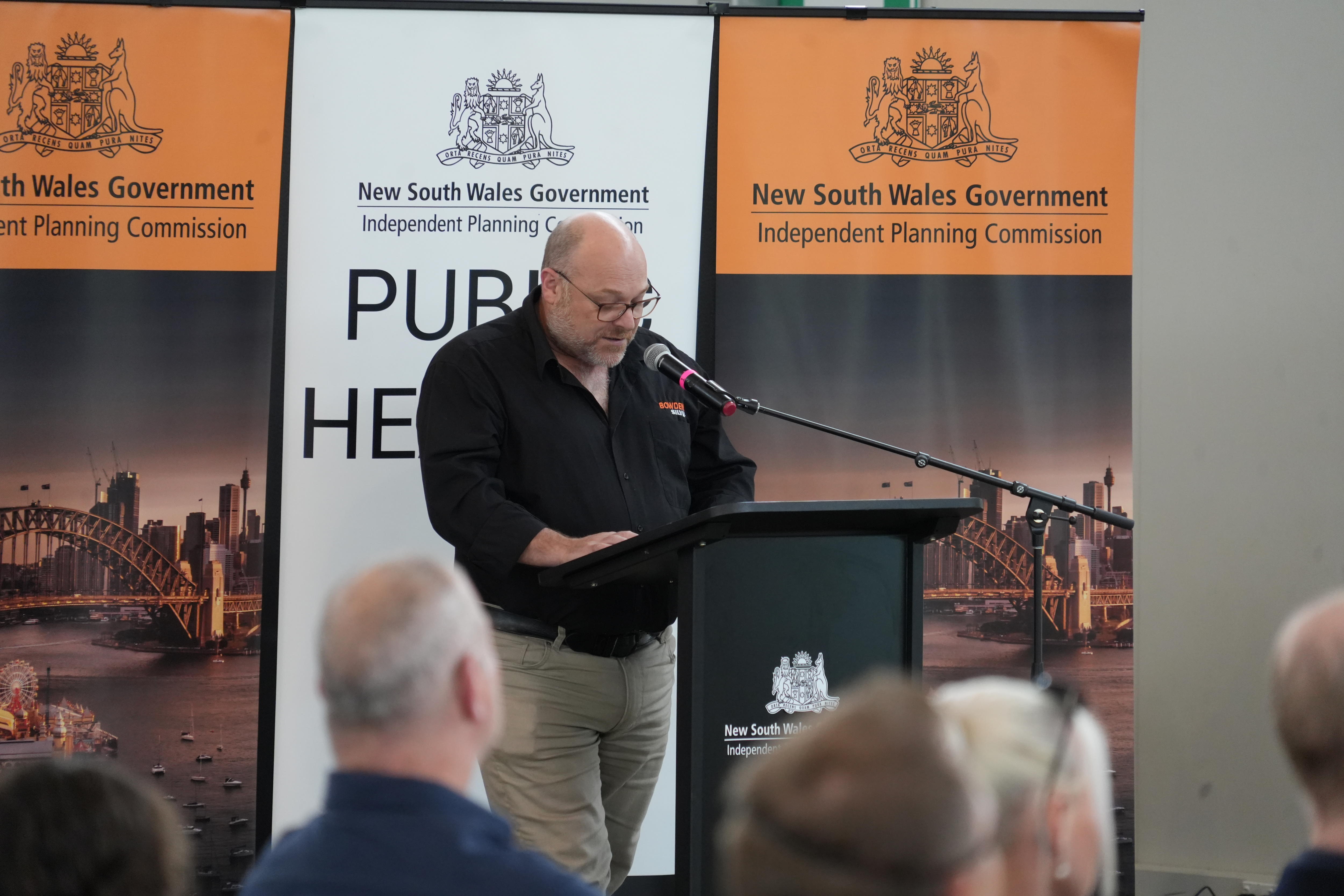 A balding man wearing glasses standing at a NSW lectern and reading from his notes.