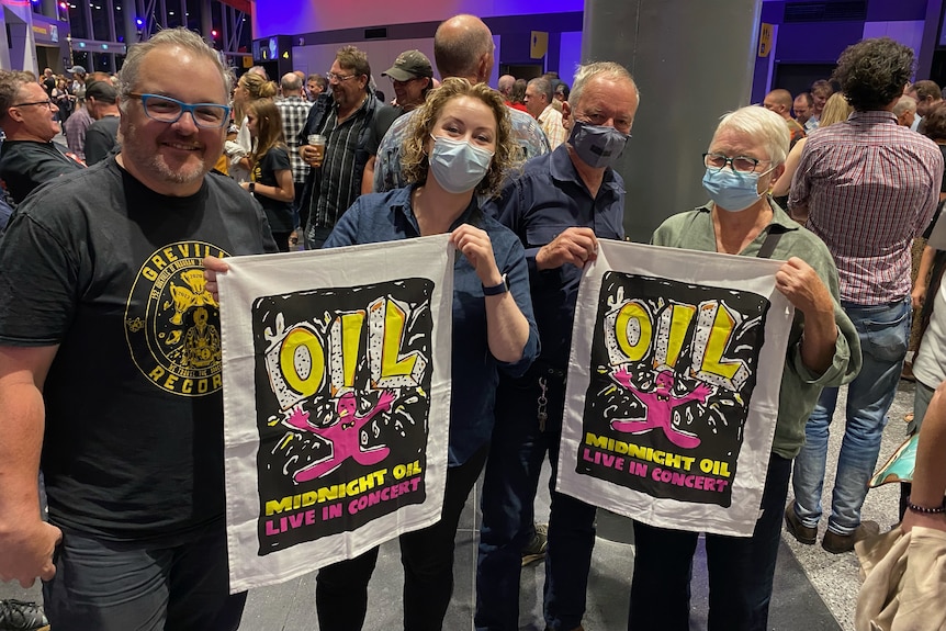 Four people, three wearing face masks, holding posters for the band Midnight Oil, with a crowd of people standing behind them.