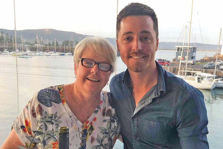 A woman in black spectacles poses for a photo by a harbour with a young man in a denim shirt