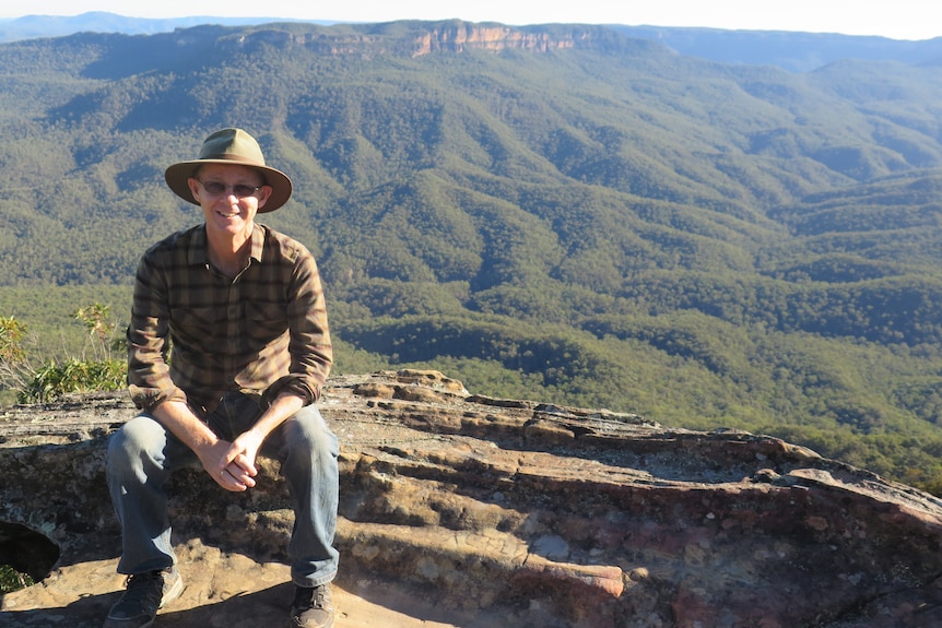 Image an older man with a wide hat on. He sits on a rocky ledge with a scenic view of mountains behind him.