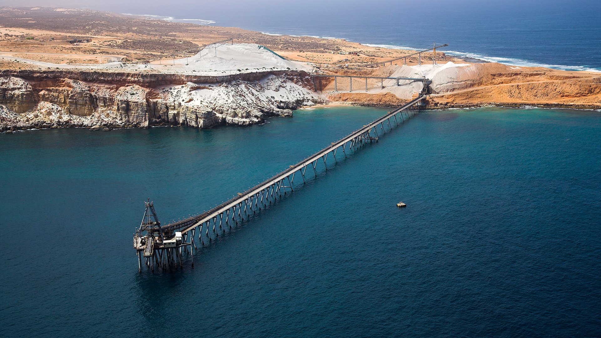 An aerial shot looking down at a long jetty with a loader off a rugged piece of orange North West coast.