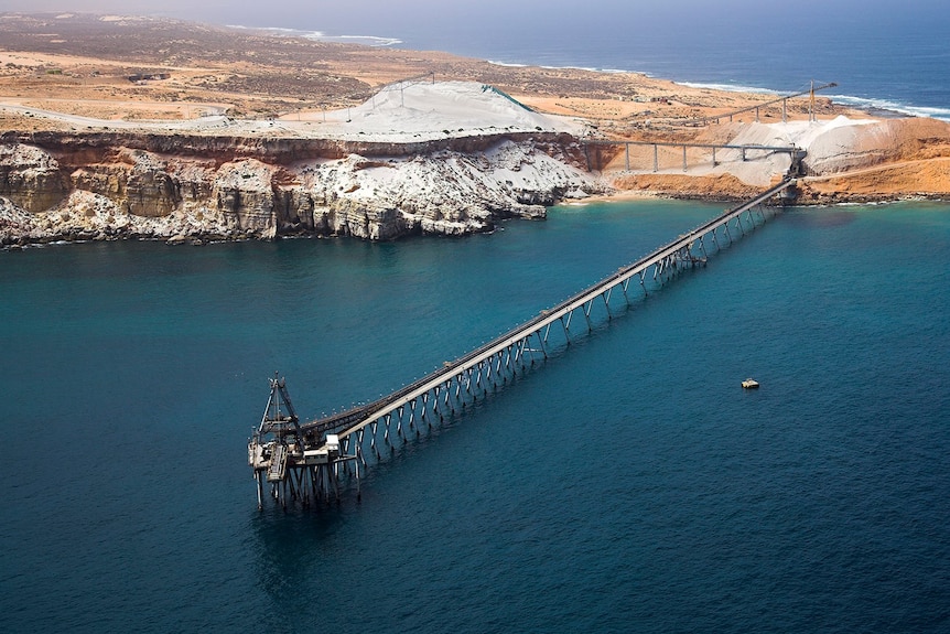 An aerial shot looking down at a long jetty with a loader off a rugged piece of orange North West coast.