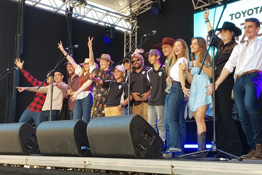 Excited young people in country music attire stand on stage in front of mic stands looking towards audience