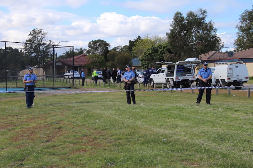 Three police officers stand on grass behind crime tape, with more on the street behind.