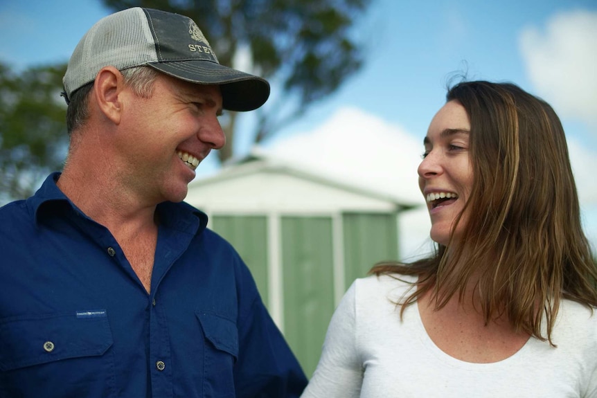 A couple smile at the camera in front of a shed