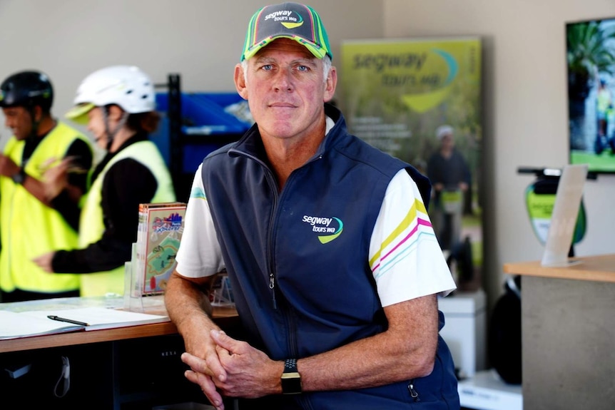 A man wearing a navy blue vest with a 'Segway Tours WA' logo stands leaning on a counter.