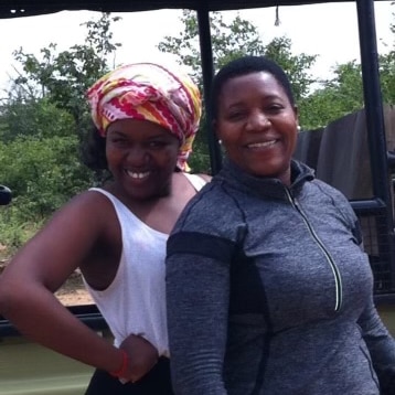 Ngwenya at age 15 wearing a white singlet and colourful head scarf, standing with her mother, a Black woman in a grey jacket