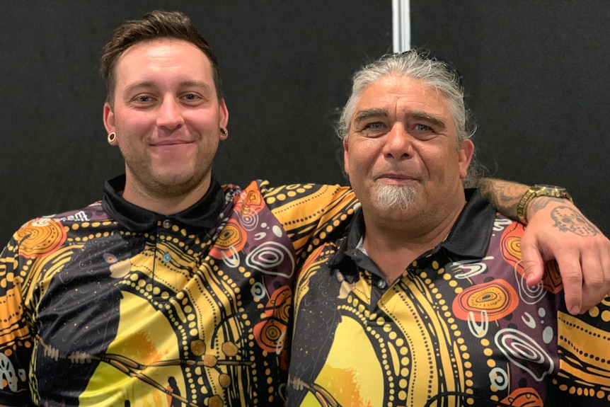 A smiling young man puts his arm around an older man. Both are wearing shirts emblazoned with an Indigenous design.