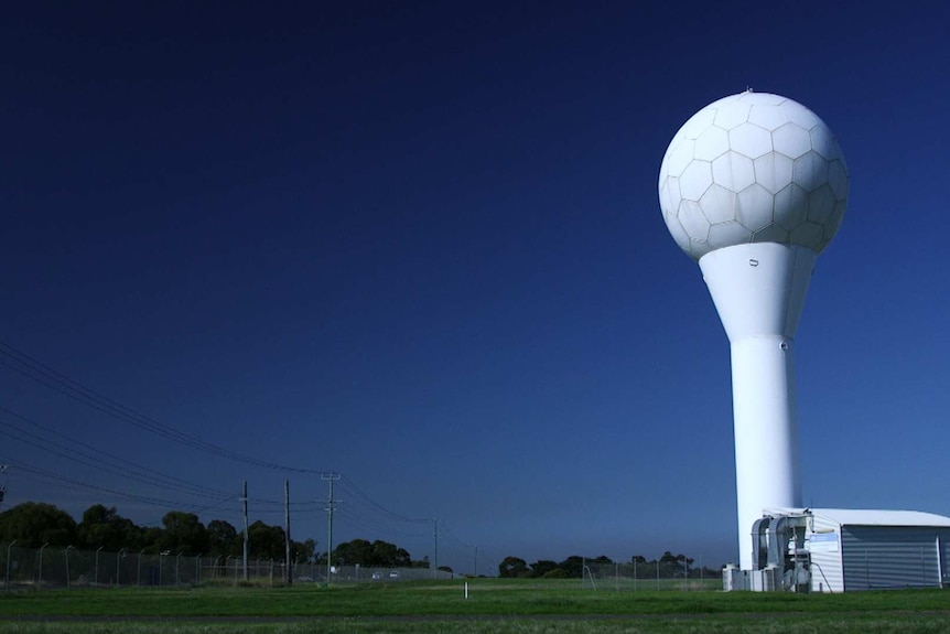 A white tower with a ball-shaped top stands next to a shed in a grassy field.