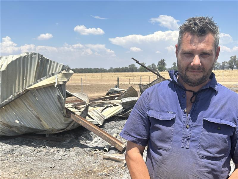 Un granjero con una camisa de trabajo azul se encuentra frente a un cobertizo arrasado en una propiedad rural.