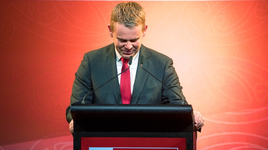 Chris Hipkins bows his head while standing at the lectern. 