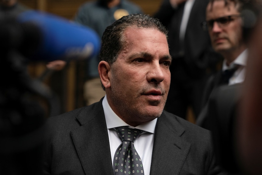 A close up of a man with short dark hair in suit and tie speaking at press conference