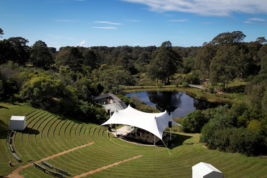 A drone shot of a sloping hill, with seating built into it and a stage at the bottom, overlooking a small lake and forest.