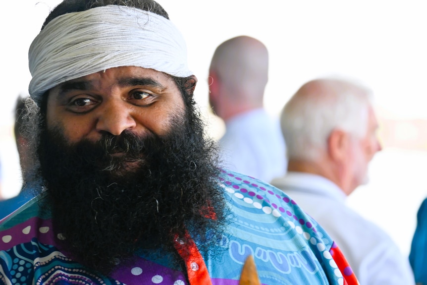 A man in colourful clothing and a white bandanna around his head and a large, black bushy beard