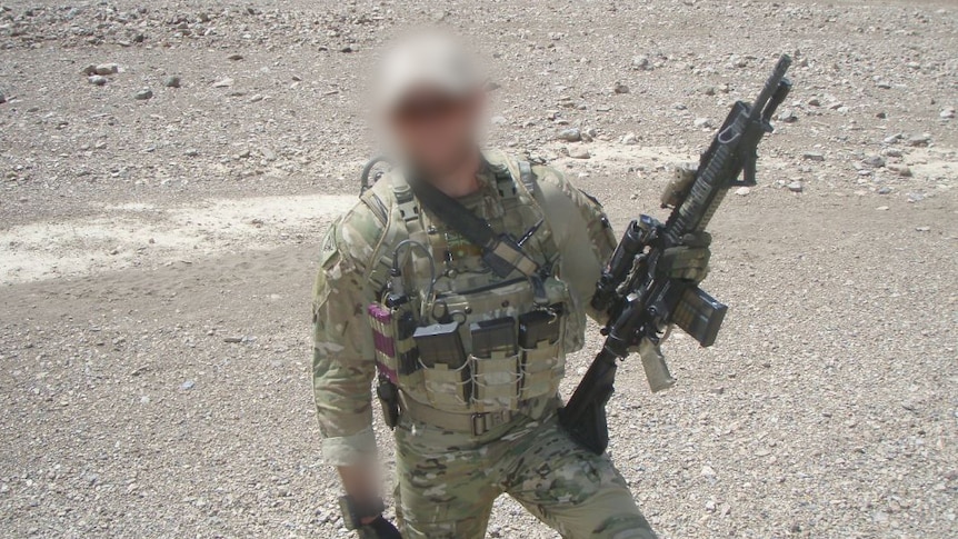 A soldier stands with a machine gun with a gravelly backdrop.
