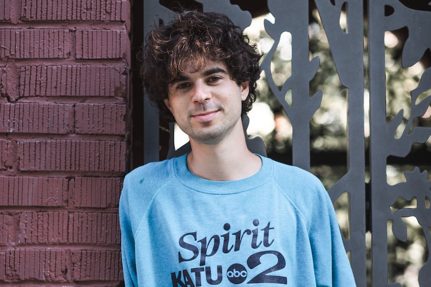 A man with curly brown hair wearing a light blue jumper leans against a brick wall.