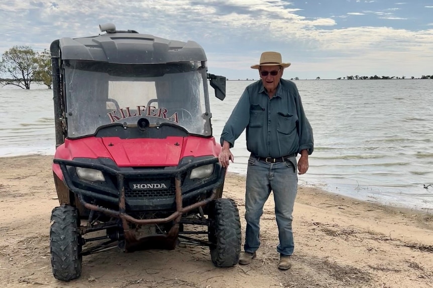 An older man wearing a hat blue shirt and jeans standing next to a red buggy on the shore of a lake