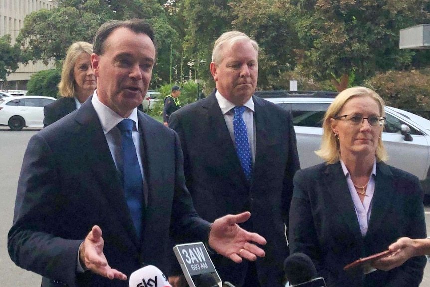 A politician standing in front of three microphones surrounded by three other people.