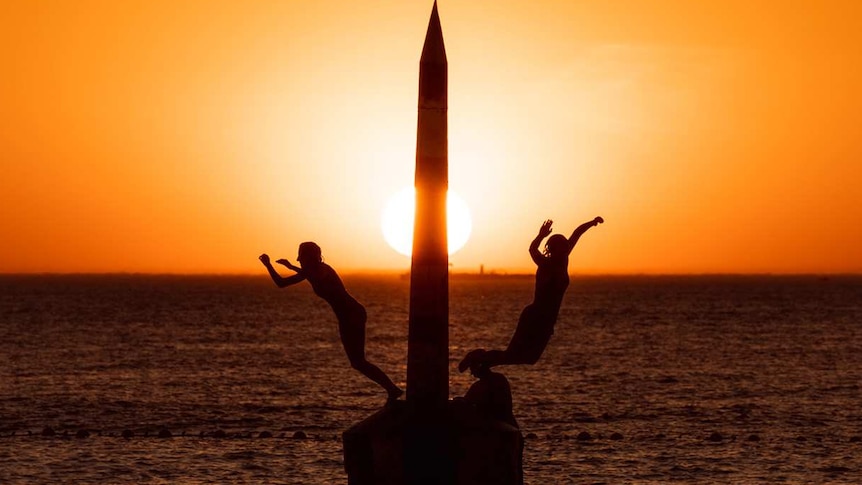 The setting sun turns the sky bright orange over the ocean at Cottesloe Beach as two people jump off a pylon into the water.