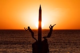 The setting sun turns the sky bright orange over the ocean at Cottesloe Beach as two people jump off a pylon into the water.