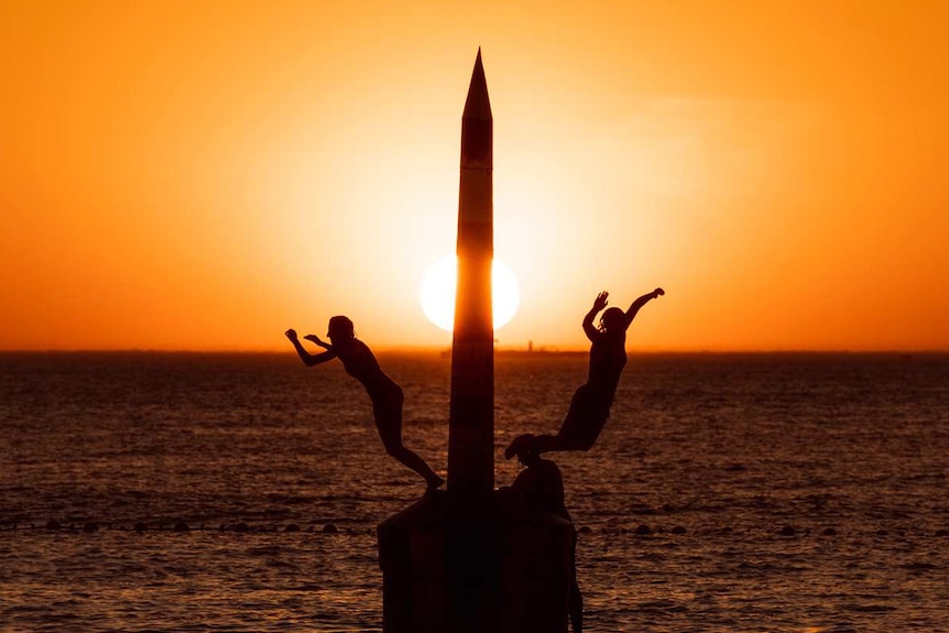 The setting sun turns the sky bright orange over the ocean at Cottesloe Beach as two people jump off a pylon into the water.