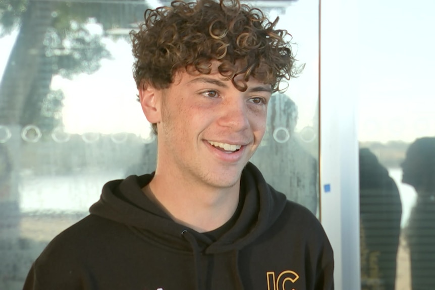 A teenage boy speaking to camera next to a glass reflective door