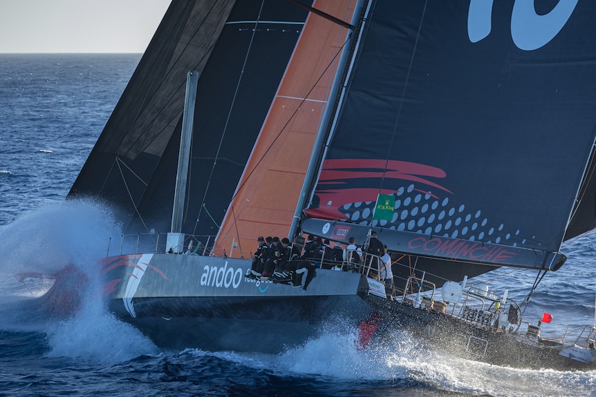 A large yacht sails on the ocean, waves splashing over the crew sitting on the side.