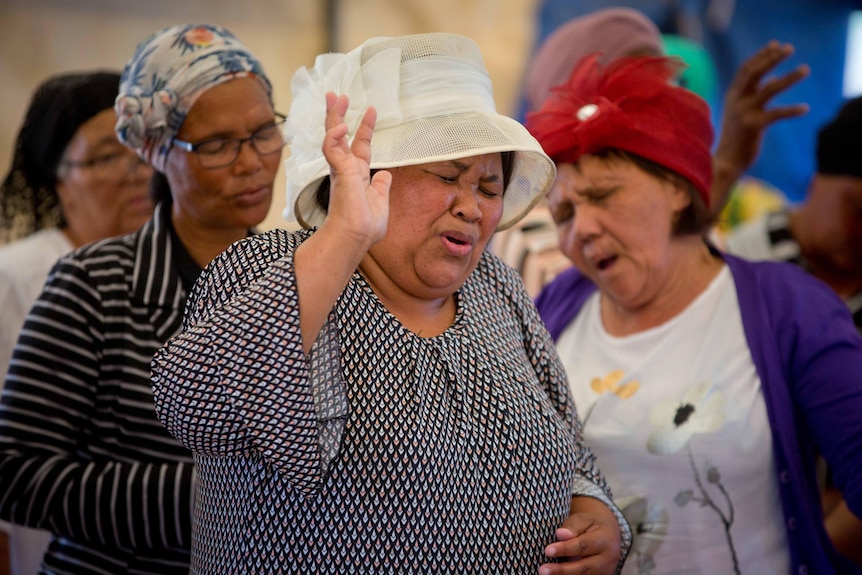 Women dressed in their Sunday best sing with their arms raised and their eyes closed.