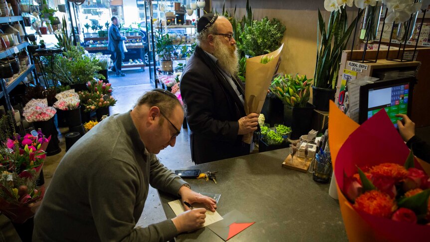 In a flower shop a man holds a bouquet while another writes a greeting card and the florist rings up another bunch of flowers.