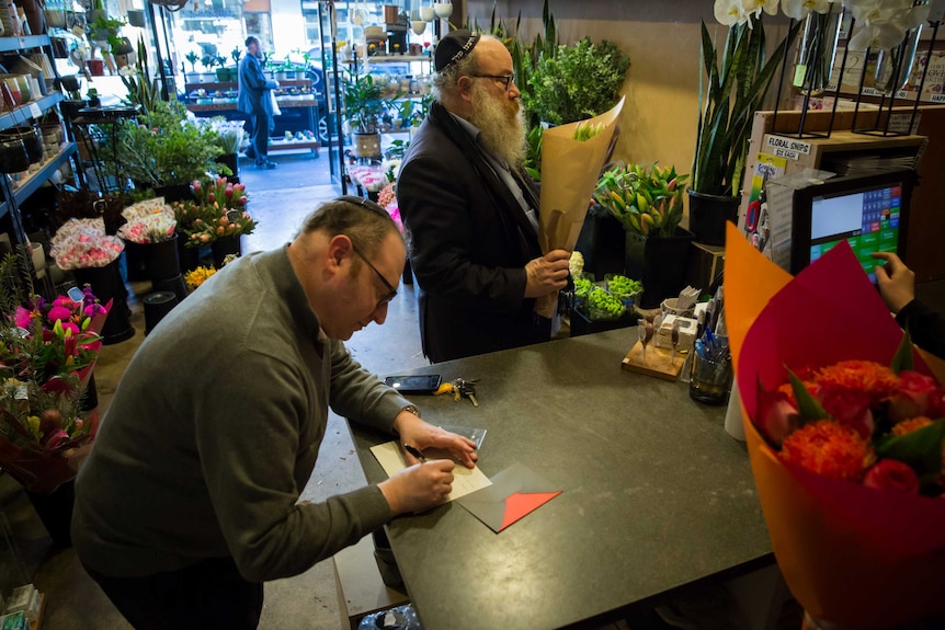 In a flower shop a man holds a bouquet while another writes a greeting card and the florist rings up another bunch of flowers.