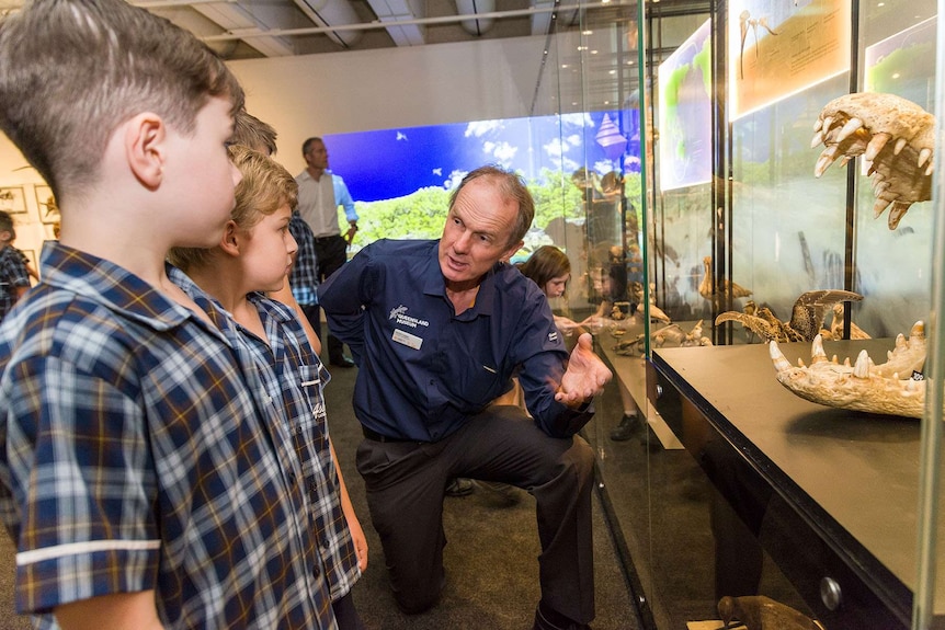 A man stands next to a case of reptile skeletons speaking to two small school boys.