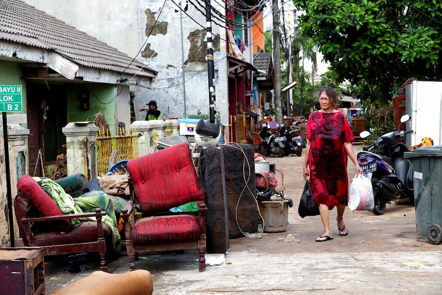 A narrow Jakarta street is pictured with debris strewn across it as a woman in red dress carries plastic bags.