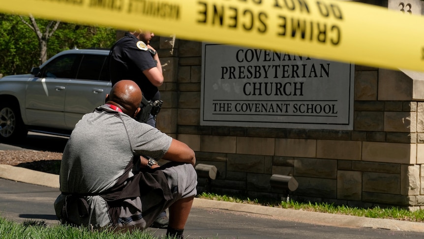 A man sits by the school sign where yellow police tape has been placed.