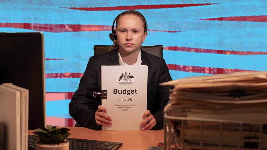 A student actor playing the treasurer sits at a desk holding bound papers with Budget on it.