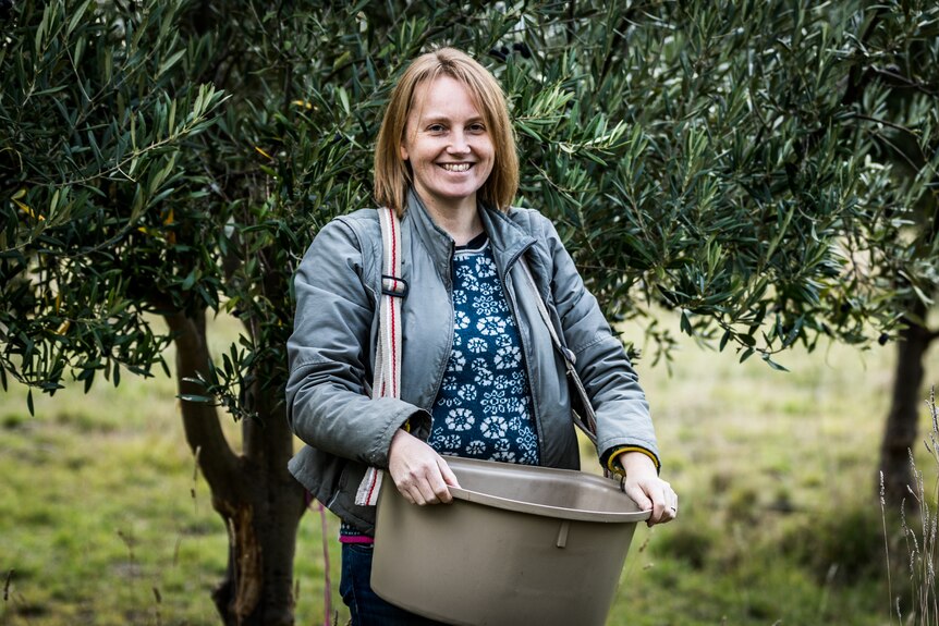 A woman stands in front of olive trees and smiles at the camera with a large picking bag strapped to her chest 