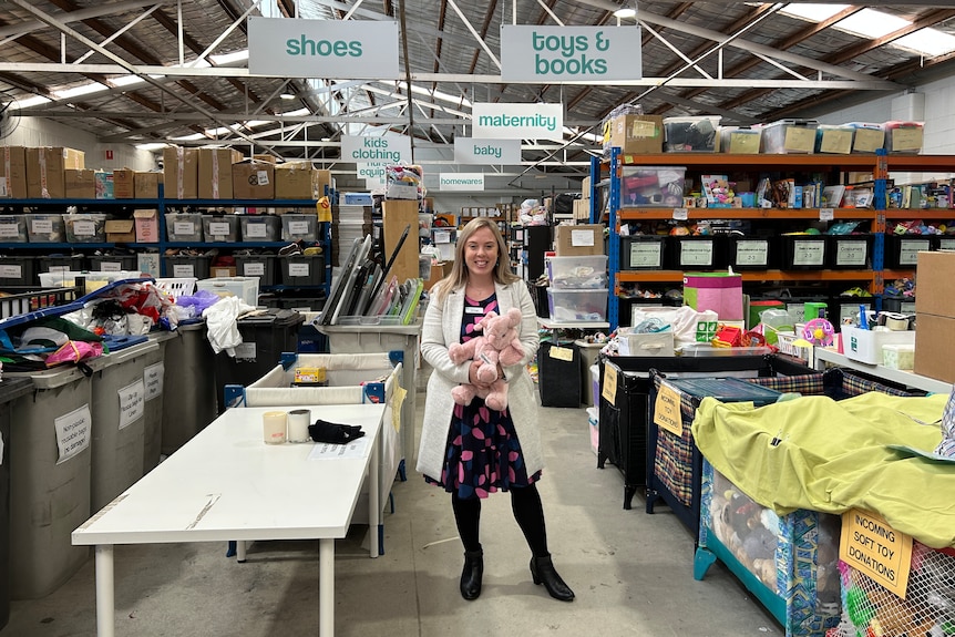 Elisa Curtis stands in a warehouse full of donated items holding a pink soft tou