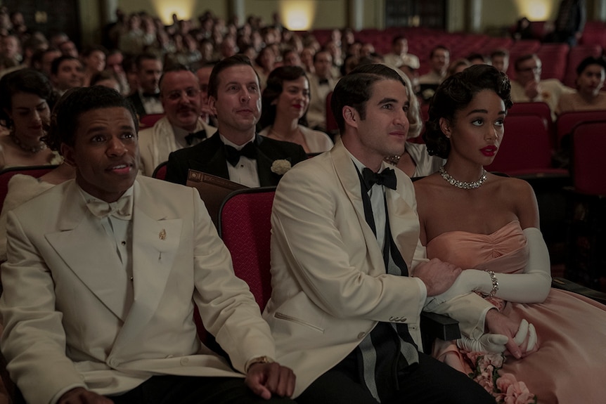 In theatre with red seats a large crowd dressed in white and black tuxedos and evening gowns look up in anticipation.