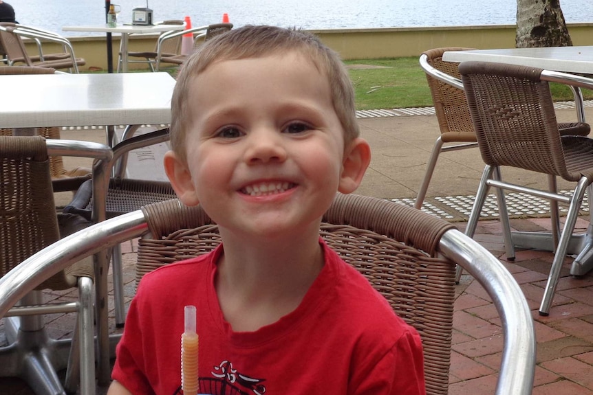 Three-year-old boy William Tyrell sits on a chair smiling holding a drink cup