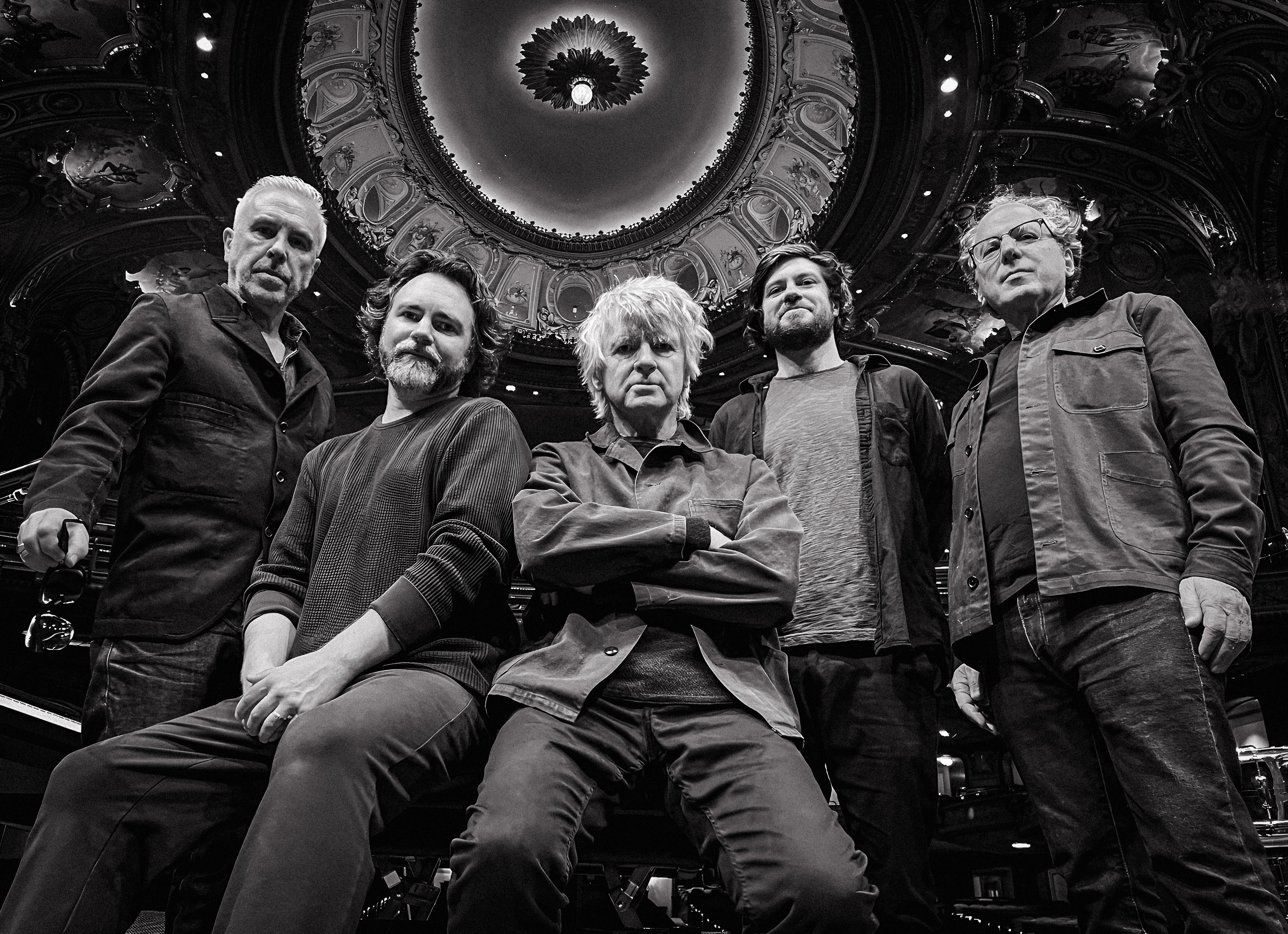 Black and white image of rock band Crowded House standing together in an auditorium with ornate ceilings
