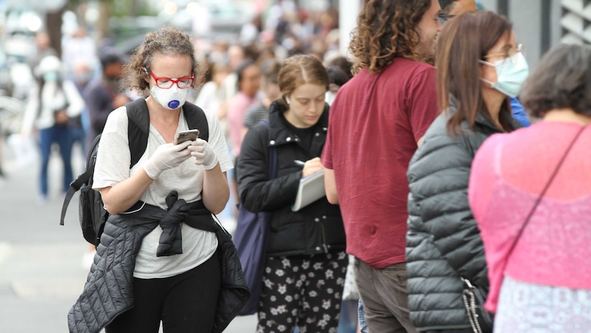 A woman wearing a mask and gloves waits in line outside Centrelink.