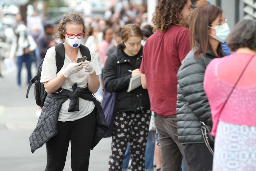 A woman wearing a mask and gloves waits in line outside Centrelink.