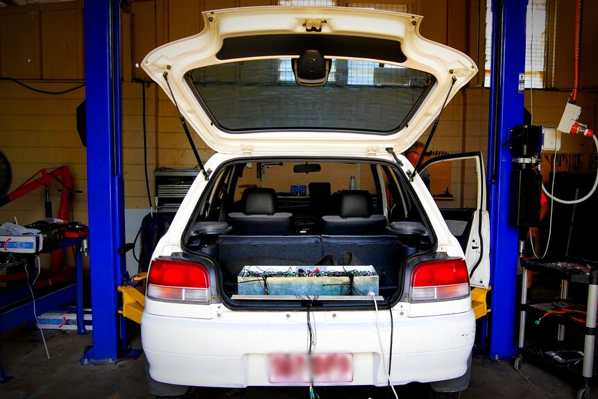 A car on a hoist in an auto-electrician's workshop, its boot open and a battery pack visible in the back.
