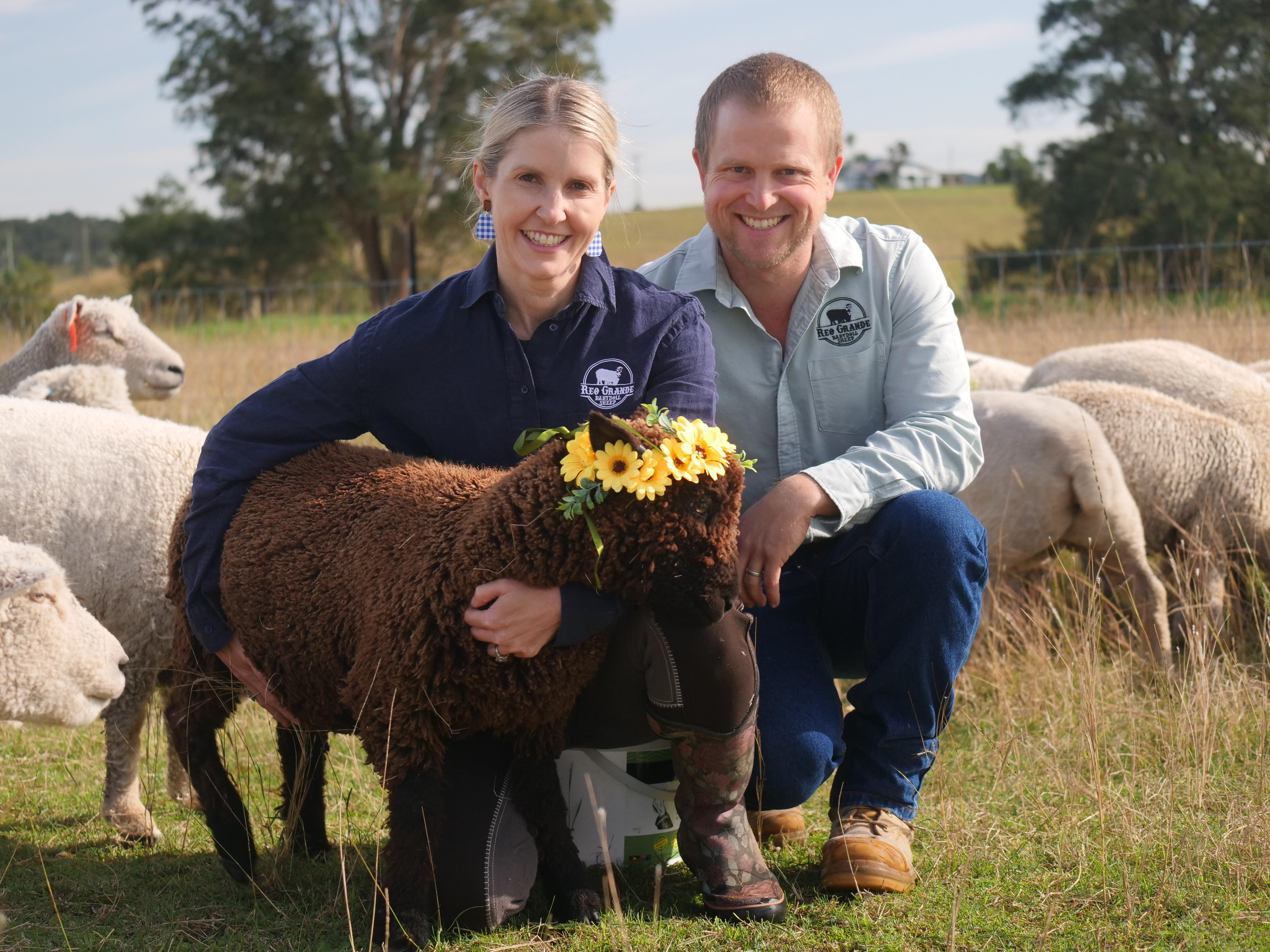 A man and woman crouch in a paddock, the woman is holding a brown babydoll sheep with a flower crown.