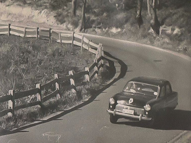 Aged black and white photo of a black car leaning with speed around a tight bend and post and rail fence.